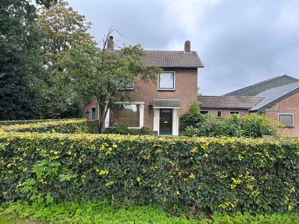 Het bakstenen woonhuis aan de Veendijk met de schuur ernaast, achter een groene haag