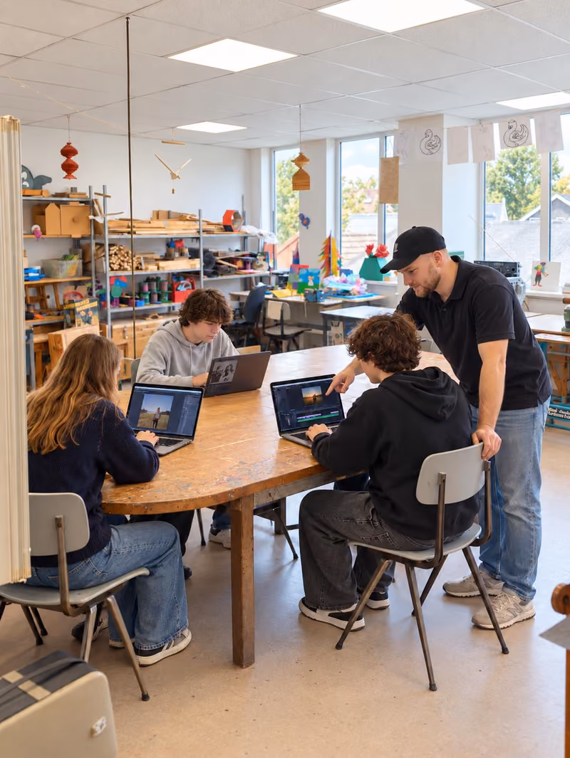 Drie jongeren werken op laptops aan een ronde tafel op De Wik, een begeleider kijkt mee