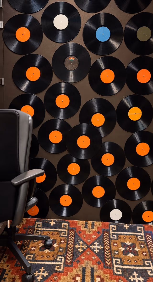 Een muur vol langspeelplaten met oranje labels in de muziekstudio, met een bureaustoel en een kleurig kleed