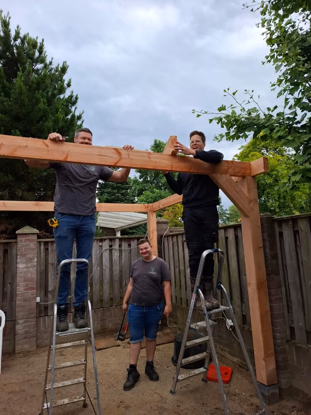 Twee begeleiders op trappen tillen een balk op een houten overkapping, een deelnemer kijkt lachend toe