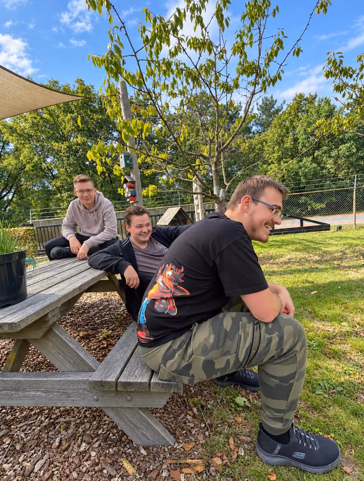 Drie deelnemers zitten lachend op en aan een picknickbank onder een schaduwdoek